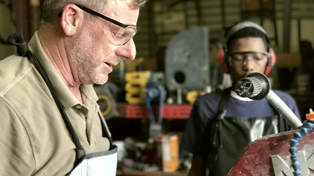 Un formateur explique un geste technique à un apprenant équipé de lunettes et d’un casque de protection, dans un atelier.