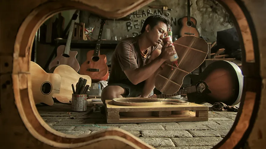 Un luthier assemble une guitare dans son atelier, vu à travers la caisse d’un instrument.